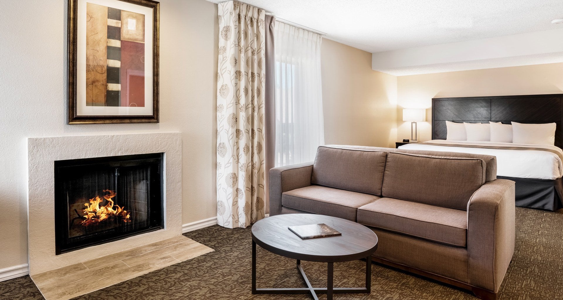 Suite living room with a lit fireplace, sofa and king bed at Chase Suite Hotel El Paso